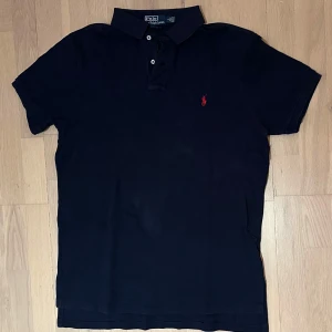 Marinblå pikétröja från Ralph Lauren - Klassisk marinblå pikétröja från Ralph Lauren med röd broderad logga på bröstet. Tröjan har korta ärmar, krage och knäppning med två knappar. Tillverkad i mjuk bomull som är skön mot huden. Perfekt för en stilren och avslappnad look.