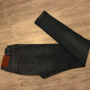 Mörkblå jeans från Tiger of Sweden - Snygga mörkblå jeans från Tiger of Sweden med klassisk femficksdesign och bruna sömmar. Jeansen har en smal passform och är tillverkade i stretchigt denim för extra komfort. Perfekta för dig som gillar en stilren och modern look.
