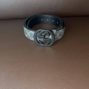 Svart och grått bälte - Snyggt bälte i svart och grått med klassiskt mönster och stort svart spänne. Bältet är brett och har en lyxig känsla, perfekt för att lyfta vilken outfit som helst. Materialet är skinn med canvasdetaljer.