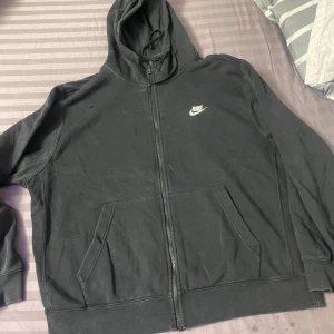 Svart Nike hoodie med dragkedja - Svart hoodie från Nike med vit logga på bröstet. Tröjan har huva med snörning, två fickor framtill och hel dragkedja. Tillverkad i mjuk bomullsblandning, perfekt för chill eller träning. Klassisk och enkel design som funkar året runt.