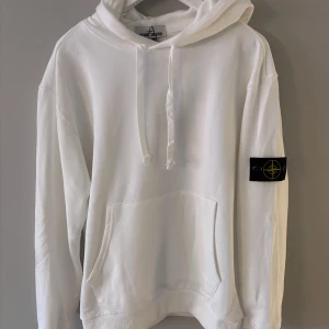 Vit hoodie från Stone Island - Säljer en snygg vit hoodie från Stone Island. Den har en klassisk design med en stor ficka fram och en justerbar huva. På ärmen finns det ikoniska Stone Island-märket. Perfekt för både vardag och chill.