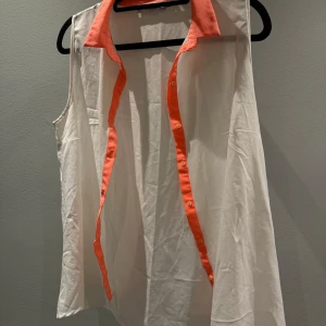 Vit ärmlös blus med korallfärgade detaljer - Säljer en snygg vit ärmlös blus med krage och knappslå i korall. Blusen har klassisk skjortkrage och knappar framtill, perfekt för en fräsch och stilren look. Materialet känns lätt och luftigt.