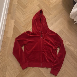 Röd velour hoodie från Juicy Couture - Juicy Couture hoodie i röd velour med dragkedja, huva och snörning. Klassisk broderad JC-logga på bröstet och känguruficka framtill. Storlek large men har blivit S/m. Köptes förra vintern knappt använd