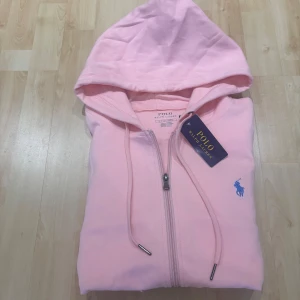 Rosa hoodie från Polo Ralph Lauren - Säljer en ljusrosa hoodie från Polo Ralph Lauren med dragkedja och huva. Tröjan har snörning i huvan, två fickor framtill och en blå broderad logga på bröstet. Tillverkad i mjuk bomull, perfekt för chill dagar.