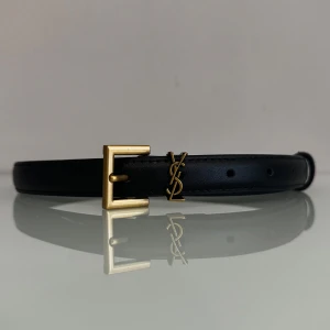 Svart skärp ysl - Snyggt svart bälte med guldig fyrkantig metallspänne och ikoniska YSL-loggan. Smal och stilren design som lyfter vilken outfit som helst. Perfekt accessoar för dig som gillar exklusiva detaljer och klassisk look. Storlek 70 (59-81)cm 