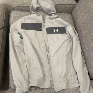 Ljusgrå vindjacka från Under Armour - Säljer en ljusgrå vindjacka från Under Armour med huva och dragkedja. Jackan har en mörkgrå panel över bröstet med logga, två sidofickor och elastiska muddar. Perfekt för blåsiga dagar och sportiga outfits.