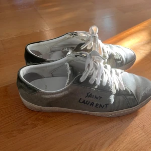Saint laurent sl06 - Gröna sneakers från Saint Laurent med vit sula och vita snören. Skorna har en kombination av canvas och mocka, med handskriven logga på sidan. Klassisk låg modell med rund tå och platt sula, perfekt för en avslappnad och trendig look. Sulan håller på att lossna lite men går enkelt att limma hos en skomakare om man vill.