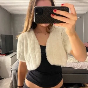 Beige bolero från Kit Karnaby - Supermjuk och fluffig beige bolero från Kit Karnaby. Den har kort modell och är fodrad med glansigt tyg på insidan. Perfekt att slänga över en klänning eller topp för en extra touch. Snygg och mysig accessoar som lyfter din outfit.