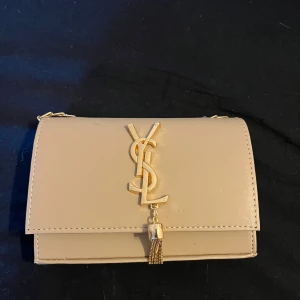 Beige YSL axelväska med guldkedja - Snygg beige axelväska från YSL med klassisk guldlogga och dekorativ tofs framtill. Väskan har en stilren, rektangulär form och kedjerem i guld. Tillverkad i slätt skinn och passar perfekt när du vill lyfta din outfit med något extra. Pris går att diskuteras 