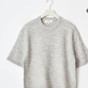 Grå stickad tröja med kort ärm GINA  - Supermjuk och stilren grå stickad tröja från ARKET med korta ärmar och rund halsringning. Perfekt för lager-på-lager och enkel att matcha med både jeans och kjol. Tröjan har en avslappnad passform och ribbade kanter för extra detalj.