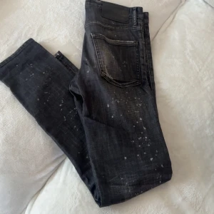 Svarta Dsquared2 jeans med dragkedja - Snygga svarta jeans från Dsquared2 med slitna detaljer, stora revor vid knäna och coola färgstänk. Tydlig silverfärgad dragkedja framtill med Dsquared2-logga. Jeansen har en smal passform och klassisk femficksdesign. Perfekta för en edgy streetstil. Extremt sällsynta och sitter som 29-30  (44 i dsq2 storlek) pris ej huggtänder i sten vid snabb affär 