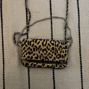 Leopardmönstrad axelväska med kedja - Väska från Zadig Voltaire. Cool axelväska med leopardmönster och silverfärgad kedjerem. Väskan har en dragkedja framtill. Perfekt för dig som vill sticka ut med en trendig accessoar.