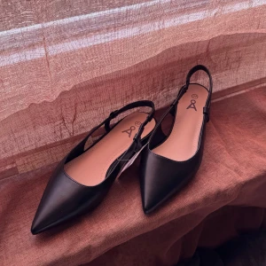 Svarta ballerinaskor/slingback med spetsig tå - Stilrena svarta ballerinaskor/slingbacks från Åhléns med spetsig tå och öppen häl. Skorna har en tunn rem runt hälen och låg klack. Aldrig använda, skulle säga att de är små i stoleken så kan passa 36/37. Köpt för 299 säljer för 200