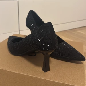 Svarta glittriga pumps med spetsig tå - Säljer ett par svarta pumps med glittriga detaljer och spetsig tå. Skorna har en låg klack och är täckta av små svarta stenar som ger en snygg glittrig effekt. De är aldrig använda. 