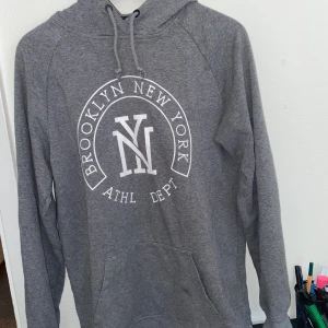 Grå hoodie Brooklyn New York - Säljer en grå hoodie med tryck 'Brooklyn New York ATHL DEPT' framtill. Tröjan har huva med snörning och en stor magficka. Perfekt för dig som gillar sportig och avslappnad stil.