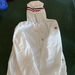 Vit vindjacka från Moncler med huva - Snygg vit vindjacka från Moncler med huva och dragkedja. Jackan har Moncler-logga på ärmen och coola ränder i rött och blått på huvan och ryggen. Tillverkad i lätt polyester och har snörning vid huvan för extra stilpoäng.