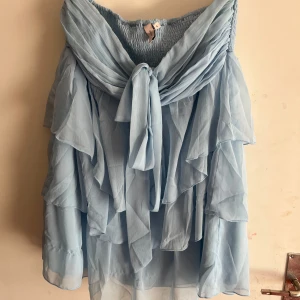 Ljusblå volangklänning från Nelly - Supersöt ljusblå kortklänning från Nelly med flera lager volanger och offshoulder-design. Klänningen har smock upptill och ett snyggt knyt framtill. Original pris 499, andvänd 1 gång