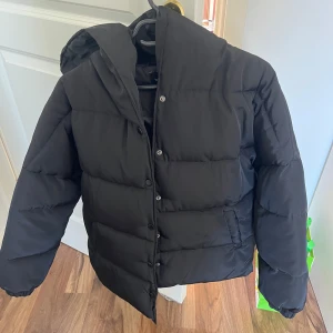 Svart jacka från Boohoo - Nypris 1200kr. Endast testad. Svart pufferjacka från Boohoo med huva och knäppning framtill. Jackan är vadderad och har två fickor på sidorna. Perfekt för kalla dagar och har en klassisk, enkel look som passar till det mesta.