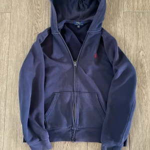 Marinblå hoodie från Polo Ralph Lauren - Säljer en marinblå hoodie från Polo Ralph Lauren med dragkedja och huva. Tröjan har det klassiska röda broderade logotypen på bröstet.