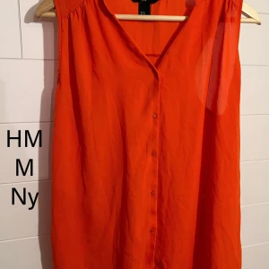 Orange ärmlös blus från H&M - Säljer en snygg orange blus från H&M i storlek M. Blusen är ärmlös med v-ringning och knappar framtill. Den har en luftig och lätt känsla, perfekt för varma dagar. Enkel och stilren design som är lätt att matcha med olika outfits.