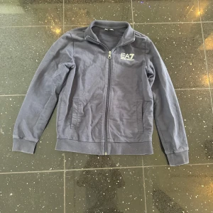Mörkblå ziptröja från EA7 Armani - Snygg mörkblå ziptröja från EA7 Emporio Armani med vit logga på bröstet. Tröjan har hel dragkedja, ståkrage och två fickor framtill. Tillverkad i mjuk bomull, perfekt för en chill och sportig look. Väldigt bra skick. 