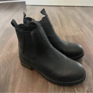 Svarta chelsea boots i skinn - Stilrena svarta chelsea boots med resår på sidorna och dragflikar fram och bak. Klassisk rund tå och låg klack, tillverkade i skinn. Perfekta för en snygg och enkel look året runt. Använda men i fint skick! 
