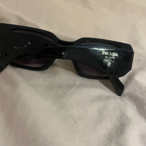 Svarta solglasögon P - Snygga svarta solglasögon P med rektangulära bågar och diskret logga på sidan. Glasögonen har en modern, kantig form och mörka linser som ger en cool vibe. Perfekta för att lyfta din stil till nästa nivå.