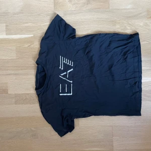 Svart EA7 t-shirt med vit logga - Svart t-shirt från EA7 Armani med vit grafisk logga på bröstet. Klassisk rund halsringning och korta ärmar. Tillverkad i mjuk bomull för en skön känsla. Perfekt för dig som gillar stilrena och sportiga plagg. Storlek S , passar 160-170 .väldigt sliten