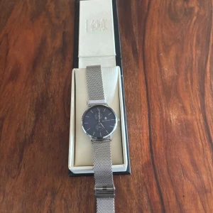 Silverfärgad klocka från Black Oak - Stilren klocka från Black Oak med svart urtavla och silverfärgat mesharmband i metall. Klockan har minimalistisk design med tunna visare och index, samt två små urtavlor. Levereras i originalask.