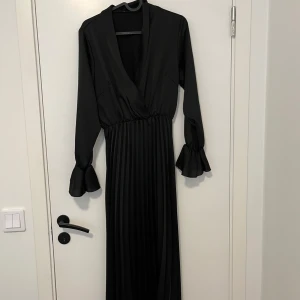 Svart långklänning med volangärm - Elegant svart långklänning med omlottliv, djup v-ringning och långa ärmar med volangdetalj vid ärmslut. Kjolen är plisserad för en snygg siluett. Perfekt för dig som vill ha en stilren och sofistikerad look.