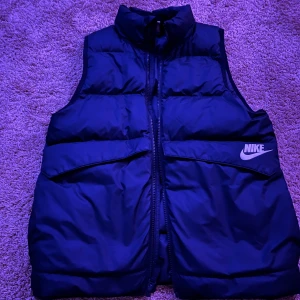 Svart dunväst från Nike, storlek 147-158cm - Svart dunväst från Nike med klassisk logga på bröstet. Västen har hög krage, två stora fickor framtill och dragkedja hela vägen. Materialet är syntet och dun, perfekt för lager-på-lager. Snygg och sportig look som funkar till streetwear. Perfekt nu till hösten att köpa till dig/ditt barn! 🍂 Använts 2 gånger sen mest hängt på en galje 😅 Snabb frakt! 🚚 