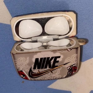 AirPods med Nike-design skal - en unikt Nike-design skal. Skalet har motiv med Nike-logga och 'Just Do It'-text, samt röda detaljer. fungerande skick. Perfekt för dig som vill sticka ut med din stil.