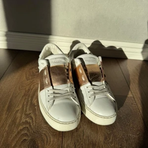 Valentino Garavani vita sneakers med guld - Snygga vita sneakers från Valentino Garavani med bred metallic guldrem över vristen och klassisk snörning. Skorna har rund tå, platt sula och är tillverkade i skinn. Perfekta för dig som vill ha en lyxig touch på din vardagsstil.