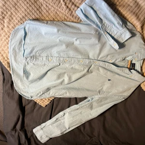 Ljusblå skjorta från Ralph Lauren - Klassisk ljusblå skjorta från Ralph Lauren med broderad logga på bröstet. Skjortan har långa ärmar, knappar framtill och är tillverkad i bomull. Perfekt för en clean och stilren look. Storleken står M men är stor i storleken.