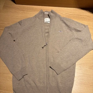 Beige kofta med dragkedja från GANT - Säljer en stilren beige kofta från GANT med hel dragkedja och liten broderad logga på bröstet. Sparsamt använd och helt felfri förutom en liten fläck som knappt syns. Perfekt för hösten och vintern, varm och otroligt bra material, köpt från NK. 