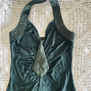 Mörkgrön halternecklänning  - Snygg mörkgrön halterneck klänning  med breda axelband som knäpps i nacken. Toppen har en markerad V-ringning och en unik sydd detalj framtill. Materialet är mjukt och har en lätt glans. Perfekt för dig som vill sticka ut med en cool och annorlunda topp. Längst ner på kjolen finns pyttesmå repor, se bild 4.