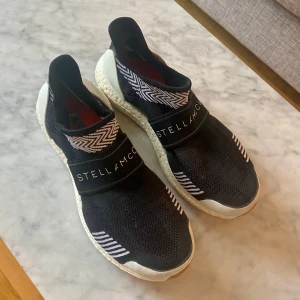 Adidas by Stella McCartney Träningsskor Sneakers 38.75 - Svarta sneakers från Adidas Stella McCartney med stickad ovandel och vita detaljer. Skorna har elastiskt band med logga över foten och mönstrad kant upptill. Sulan är vit och ger en sportig look. Perfekta för dig som gillar stilrena och bekväma sneakers.