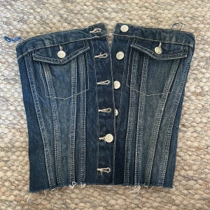 Jeanskorsett med knappar och fickor - Säljer en snygg korsettopp i denim med silverfärgade knappar framtill och två dekorativa bröstfickor. Toppen har markerade sömmar, rå kant nertill och elastisk smock baktill för skön passform. Perfekt för dig som vill sticka ut med en cool jeanslook.