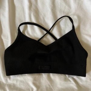 Svart ribbad linnetopp Björn Borg Alice stenlöf  - Svart ribbad linnetopp från Björn Borg x Alice Stenlöf med smala korsade axelband. Toppen är ärmlös och har en tight passform, perfekt för träning eller chill. Diskret logga framtill och mjukt stretchigt material.