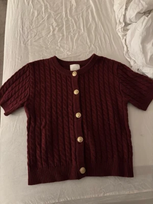 Vinröd kabelstickad kofta med kort ärm - Kortärmad vinröd kofta med kabelstickat mönster och fem guldknappar framtill. Tröjan har rund halsringning och ribbade muddar vid ärmslut och nederkant. Perfekt för lager på lager och ger en snygg retrovibe.