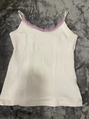 Vit linnetopp med spetskant - Söt vit linnetopp med tunna axelband och ljuslila spetsdetalj längs urringningen. Toppen är ärmlös och har en enkel, rak passform. Perfekt för sommardagar eller som lager under en skjorta.