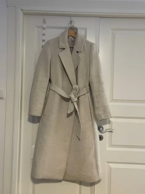 Beige kappa med bälte från H&M - Säljer en stilren beige kappa från H&M i storlek 38. Kappan har klassisk krage, knytskärp i midjan och två fickor framtill. Fodrad insida och långärmad modell som passar perfekt till kyligare dagar. Enkel och tidlös design som är lätt att matcha.