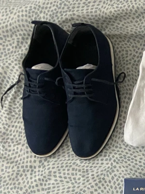 Mörkblå mode skor med vit sula - Säljer ett par stilrena mörkblå old money skor med snörning och vit sula. Skorna har en rund tå och är tillverkade i textilmaterial, vilket gör dem lätta och bekväma. Perfekta för dig som vill ha en clean och modern look. Storlek 41 men passar också för dem med strl 42.