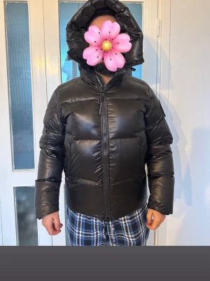 Svart pufferjacka från Canada Goose - Säljer en svart pufferjacka från Canada Goose med huva och två reflekterande ränder bak. Jackan har dragkedja framtill och patch med logga på ärmen. Materialet är glansigt och fylligt, perfekt för kalla dagar. Snygg och enkel design som håller dig varm. 