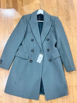 Grön kappa från Mango, dubbelknäppt - Snygg grå kappa från Mango i klassisk modell med dubbelknäppning och breda slag. Kappan har dekorativa knappar framtill och på ärmsluten samt två fickor med lock. Perfekt för dig som vill ha en stilren och tidlös look.
