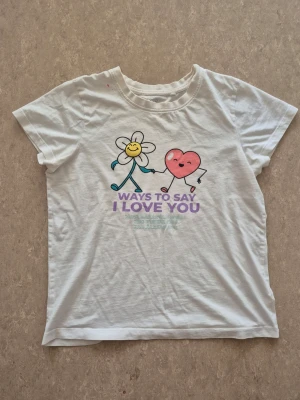 T-shirt - Vit t-shirt från Wow i bomull med korta ärmar och ett gulligt tryck framtill av en blomma och ett hjärta som håller hand. Texten 'Ways to say I love you' i lila och fler språk under. Perfekt för dig som gillar söta motiv och sköna basplagg. Finns en rosa fläck.