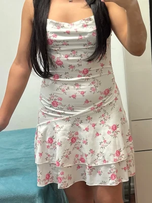 Vit blommig kortklänning med volang - Supersöt vit kortklänning med rosa blommor och volangkant. Klänningen är ärmlös och har en lätt draperad urringning. Materialet känns mjukt och stretchigt, perfekt för varma dagar. Passformen är figurnära och kjolen har dubbla lager för extra flow.