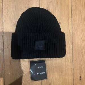   Acne Studios mössa - Säljer en svart ribbstickad mössa från Acne Studios med det klassiska ansiktslogotypen framtill. Mössan är i tjock ull och har en bred uppvikt kant för extra stilpoäng. Perfekt accessoar för kalla dagar.