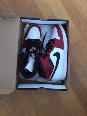 Nike Air Jordan 1 Mid röd/vit/svart 44 - Säljer ett par Nike Air Jordan 1 Mid i klassisk röd, vit och svart färg. Skorna har svart snörning, vit tåbox med perforeringar och ikonisk svart swoosh på sidan. Tillverkade i skinn och syntetmaterial, med hög siluett och röd yttersula. Perfekta för dig som gillar streetstyle.