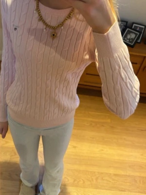 Ljusrosa kabelstickad tröja Gant - Supersöt ljusrosa kabelstickad tröja från Gant med rund hals och diskret logga på bröstet. Tröjan har lång ärm och ribbade muddar vid ärmslut och nederkant. Perfekt för dig som vill ha lite finare tröja.